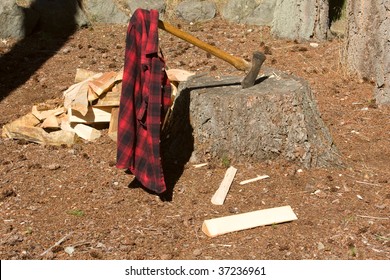 Double Bit Axe In Stump With Red Black Flannel Shirt Hanging On Handle. A Pile Of Wood Is Next To The Stump.