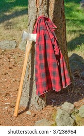 Double Bit Axe In Stump With Red Black Flannel Shirt Hanging On Handle. A Pile Of Wood Is Next To The Stump.