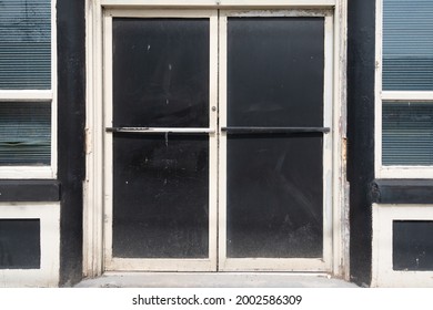 Double commercial exterior doors of a store. The glass has been painted black, trim is black and beige. The old doors have metal handles. There are large glass windows on both sides of the doors. 