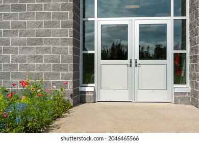 Double Commercial Exterior Steel Doors Of A Business. The Glass Is On Top, Trim Is Stainless Steel Metal. The Doors Have Metal Handles. There Are Large Grey Brick Walls On Both Sides Of The Doors. 