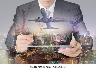 Double Exposure Of Businessman Working With Tablet, Airplane, Construction Crane And Night Cityscape As Business, Technology, Industry, Transportation And Developing Country Concept.