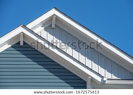 Double gable, with white decorative corbel, bracket, brace on a triangle gable roof, white soffit and fascia,  gray vertical and blue horizontal vinyl lap siding with blue sky background