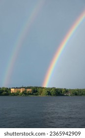 Double rainbow over water by Manilla school building by green forest on Djurgården in Stockholm