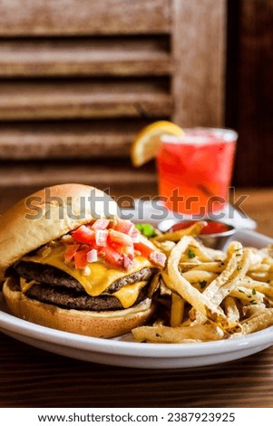 A double stack juicy cheeseburger on a toasted bun topped with tomato salsa with shoestring french fries topped with fresh herbs with a wooden chair and pink cocktail in the background.