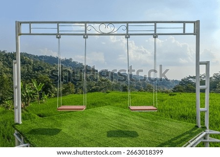 A double swing set on a platform of artificial turf overlooks a vibrant green rice field and lush, forested hills under a blue sky with scattered clouds.