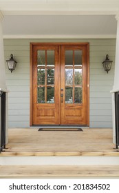 Double Wooden Doors To A Green Home. The Doors Have Windows, And Are Flanked By Twin Light Fixtures. Also Seen Is A Doormat, And A Wooden Porch.