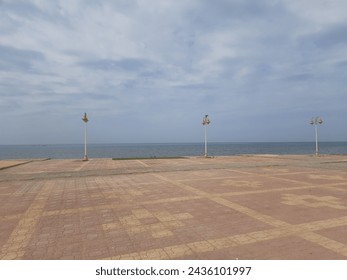 Double-bulb electric lighting poles on a red and yellow tiled cornice on the shore of the Red Sea in the city of Qunfudhah during the day when there are many clouds in the sky