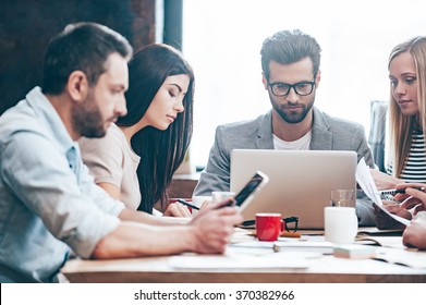 Double-checking All The Documents. Group Of Four Young People Reading And Looking Through Charts While Sitting At The Table In Office