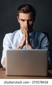 Double-checking his work. Confident handsome young man looking at his laptop and keeping handsclasped while sitting against black background