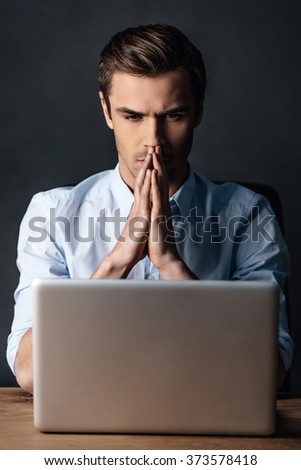 Double-checking his work. Confident handsome young man looking at his laptop and keeping handsclasped while sitting against black background