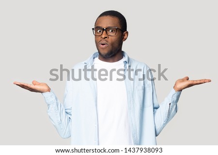Doubtful african American millennial male in glasses isolated on grey studio background compare offers or prices, unsure black man keep hands aside doubt over deal, make decision or choice