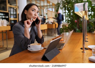Doubtful Asian young adult business woman talking on cell phone making call working looking away thinking of solution sitting in cafe, considering offer, making decision or feeling doubt. - Shutterstock ID 2189886997