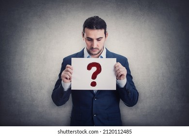 Doubtful Businessman Holding A Paper Sheet With Question Symbol, Isolated On Grey Wall Background. Depressed Person Looking Down Pensive. Interrogation Mark As Metaphor For Business Troubles