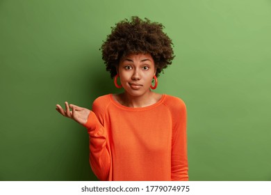 Doubtful indecisive woman raises palm with hesitation, faces difficult question or two choices, wears orange sweater and earrings, isolated on green background. People, perception and attitude