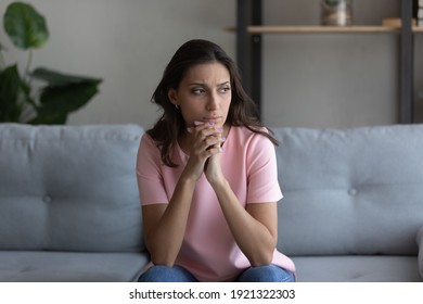 Doubtful millennial arab female sitting on couch at home lost in serious thoughts. Thoughtful young woman of indian ethnicity looking aside pondering on making choice decision solving problem in mind - Shutterstock ID 1921322303