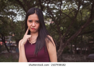 A doubtful and skeptical Filipina woman in a maroon sleeveless blouse at the park.