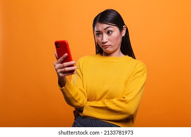 Doubtful uncertain beautiful woman wearing yellow sweater while using smartphone device on orange background. Confused clueless adult person using mobile cellphone to understand un clear instructions. - Shutterstock ID 2133781973
