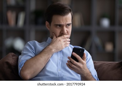 Doubtful Young Man Sit On Sofa Hold Phone Look Aside Hesitate In Making Important Choice Decision Before Make Call. Worried Concerned Millennial Businessman Think On Disturbing News Received By Email