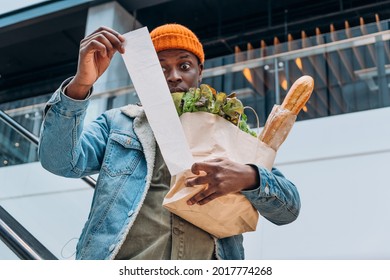 Doubting African-American person in denim jacket looks at sales paper receipt total holding pack with food products on escalator - Shutterstock ID 2017774268