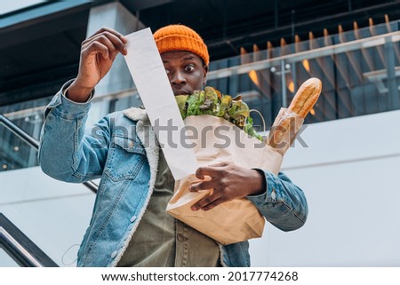 Doubting African-American person in denim jacket looks at sales paper receipt total holding pack with food products on escalator