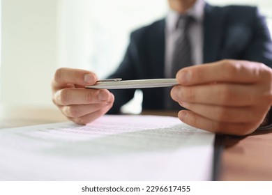 Doubting businessman holds ball pen in hands above contract papers sitting at table in office. Man studies rules of agreement before signing document