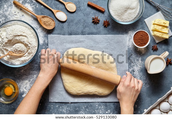 Dough\
bread, pizza or pie recipe homemade preparation. Female baker hands\
rolling dough with pin. Food ingridients flat lay on kitchen table.\
Working with pastry or bakery cooking. Top\
view