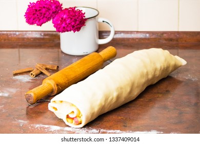 The dough is rolled into a roll on the kitchen table for making Austrian traditional apple strudel. Near the wooden rolling pin and cinnamon In the background are pink flowers in a metal cup and an ap