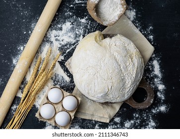 Dough sprinkled with flour, rolling pin and ears of wheat on a black table. Raw eggs, flour in a canvas bag in the background. The table is strewn with flour. Top view