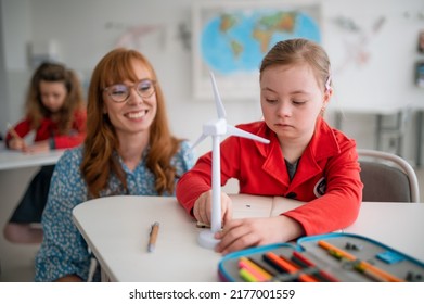 Down syndrome schoolgirl with model of wind turbine with help of teacher learning about eco-friendly renewable sources of energy in class at school, integration concept. - Powered by Shutterstock