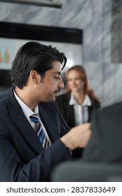 Downcast gaze from the dark-haired professional processing a problem in the marble boardroom, red-haired businesswoman soft and calm behind his shoulder in a bright modern coworking office.