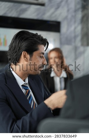 Downcast gaze from the dark-haired professional processing a problem in the marble boardroom, red-haired businesswoman soft and calm behind his shoulder in a bright modern coworking office.