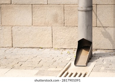 Downspout pipe of gutter system directing rainwater to sidewalk drain near building stone facade.