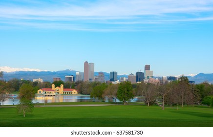 Downtown Denver skyline buildings, on a bright clear summer morning with lake and trees in foreground and snowcapped mountains in background.