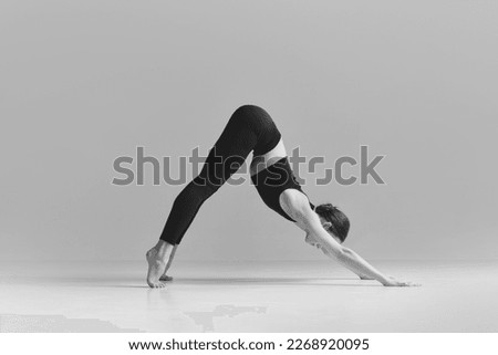 Downward facing dog pose. Black and white photography. Young girl doing stretching exercises over studio background. Concept of sport, body care, beauty, fitness, active lifestyle. Ad