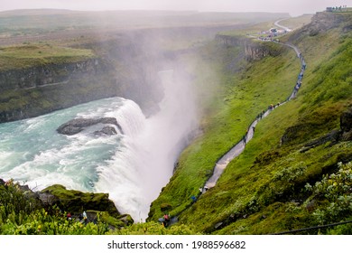 Downwards View At Gullfoss Falls Turquoise Water On A Rainy Day. Tourists' Trail Facing The Falls, By The Water Daw. Cloudy Sky And Black Volcanic Soil Canyon. Golden Circle Tour, Iceland