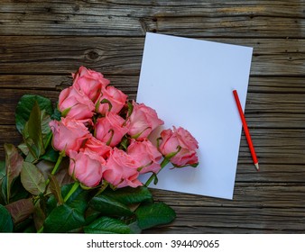 Dozen Pink Roses On Stems Beside Plain White Paper And Red Colored Pencil On A Knotted Dark Wood Table