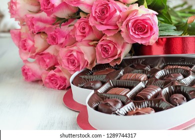 Dozen Soft Pink Rose Flowers With A Heart Shaped Box Of Chocolate Candy For Valentine Day Over A Wood Background.