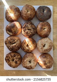 A Dozen Variety Bagels On A White Cutting Board On A Butcher Block Table Shot From Above With A Ray Of Sun Diagonally Covering The Bagels