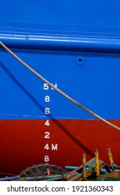 Draft Mark Numbers On Blue And Red Rust Proof Vessel Hull With Mooring Rope And Rusty Metal Scraps Pile At Shipyard In Vertical Frame, Maintenance And Improvement Concept