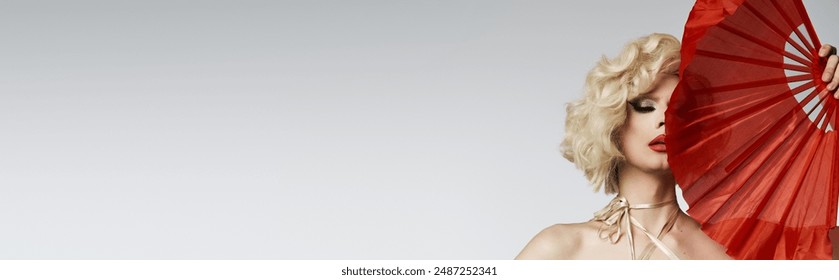 A drag queen with a blonde wig and bold makeup poses with a red fan in front of a gray background. - Powered by Shutterstock