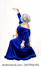 A drag queen dressed in a blue dress is sitting comfortably in a chair, exuding confidence and poise. She appears elegant and stylish in her attire.