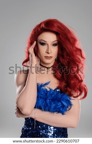 Drag queen in red wig and dress posing and looking at camera isolated on grey