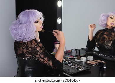 Drag Queen In Violet Wig Applying Face Powder With Cosmetic Brush Near Mirror