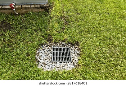 Drainage catch basin grate surrounded by decorative gravel and green grass next to a wooden platform supporting an air conditioning unit
