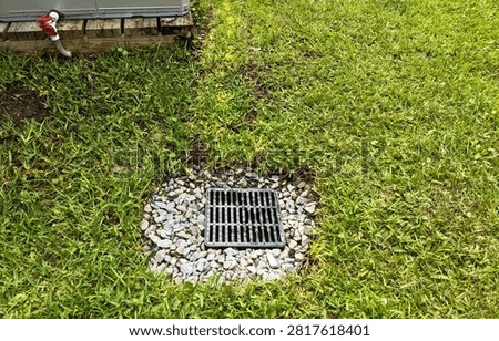 Drainage catch basin grate surrounded by decorative gravel and green grass next to a wooden platform supporting an air conditioning unit