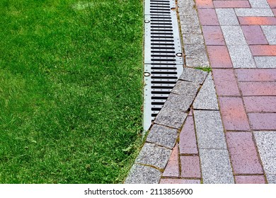 Drainage Grate Bolted To Storm Drain At Corner Of Pedestrian Pavement Walk Way Made Of Stone Brick Tiles Gray And Red Pattern In Backyard With Copy Space On Green Grass On Park, Nobody.