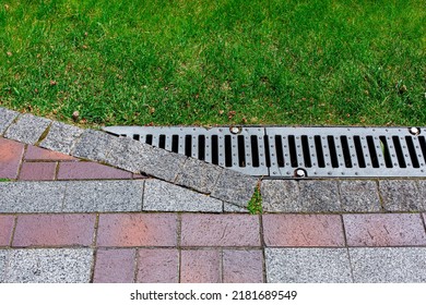 Drainage Grate Bolted To Storm Drain At Corner Of Pavement Walkway Path Made Of Stone Brick Tiles Red And Gray Color With Pattern In Backyard With Grass Close-up Of Engineering Structures Of Garden.