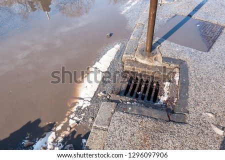 Drainage or sewage manhole grid system on the street asphalt road useless because of damaged tarmac with pot hole full of water after heavy rain and melted snow in the winter season