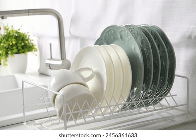 Drainer with different clean dishware and cups on white table in kitchen