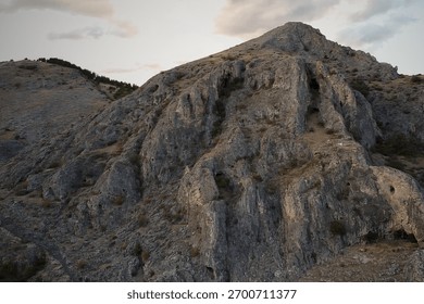 A dramatic 4K drone view of a steep, rugged mountain face. The detailed rock texture and sheer size of the cliffside dominate the frame, conveying raw natural power under a soft, cloudy sky.
